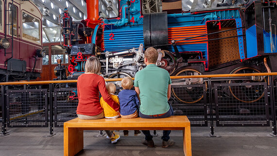 Eine Familie sitzt auf einer Holzbank und bewundert eine farbenfrohe, vintage Dampflokomotive, die längs aufgeschnitten wurde.
