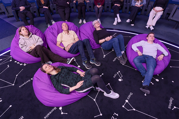 Ein gemütlicher Innenraum des Planetariums mit Menschen, die sich auf grossen lilafarbenen Sitzsäcken entspannen, umgeben von einem sternenübersäten Boden, auf dem Sternbilder zu sehen sind.