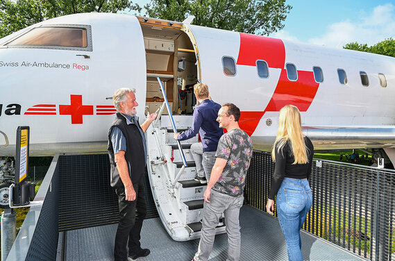 Eine Gruppe von vier Personen nähert sich einem Rega-Flugzeug und lässt sich von einem Guide durch das Flugzeug führen.