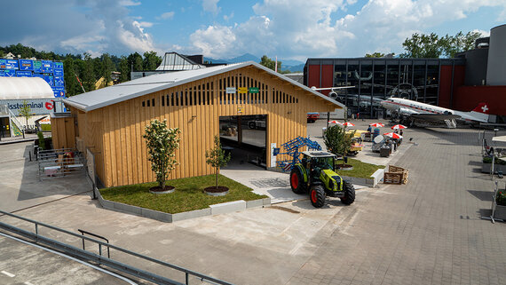 Ein Bauernhaus aus Holz mit einem Solardach und einem grünen Traktor davor steht im Zentrum der Verkehrshaus-Arena.