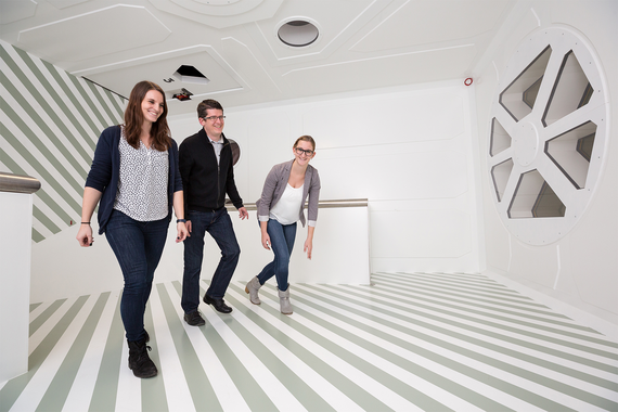 Drei Personen gehen durch ein modernes, minimalistisches Interieur mit gestreiften Fußböden und einem kreisförmigen Fensterdesign.