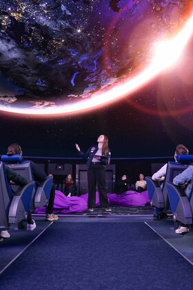 Eine Gruppe von Menschen sitzt in einem Planetarium und blickt auf eine lebendige Projektion der Erde und des Kosmos darüber.