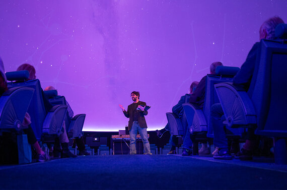Ein Moderator steht vor einem Publikum, das von einem violetten Sternenhimmel in einem Planetarium angestrahlt wird.