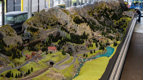 Eine detaillierte Modelllandschaft der Gotthardbahn mit Hügeln, Wäldern, gewundenen Straßen, Miniaturgebäuden und Eisenbahnschienen.