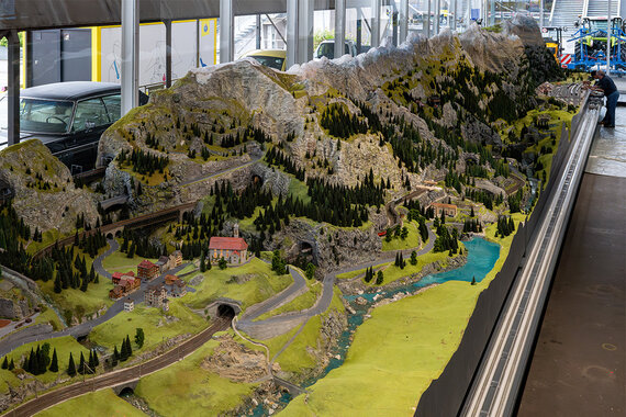 Eine detaillierte Modelllandschaft der Gotthardbahn mit Hügeln, Wäldern, gewundenen Straßen, Miniaturgebäuden und Eisenbahnschienen.