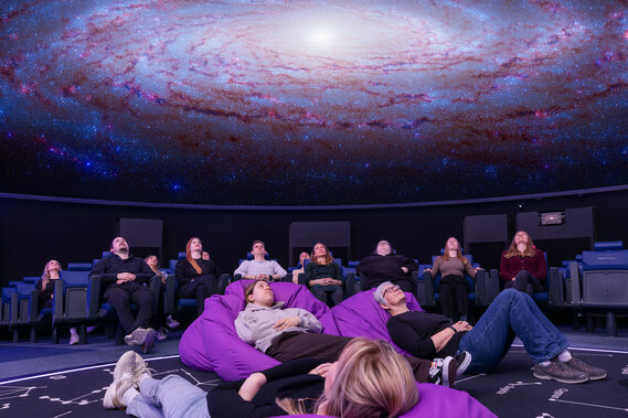 Personen sitzen in Lehnstühlen oder liegen in Sitzsäcken im Verkehrshaus Planetarium und schauen das Weltall an, das in der Kuppel projiziert wird. 