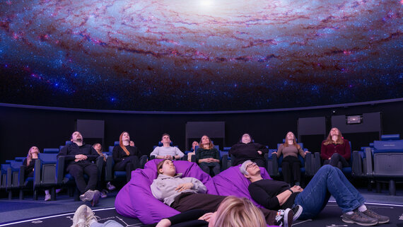 Personen sitzen in Lehnstühlen oder liegen in Sitzsäcken im Verkehrshaus Planetarium und schauen das Weltall an, das in der Kuppel projiziert wird. 