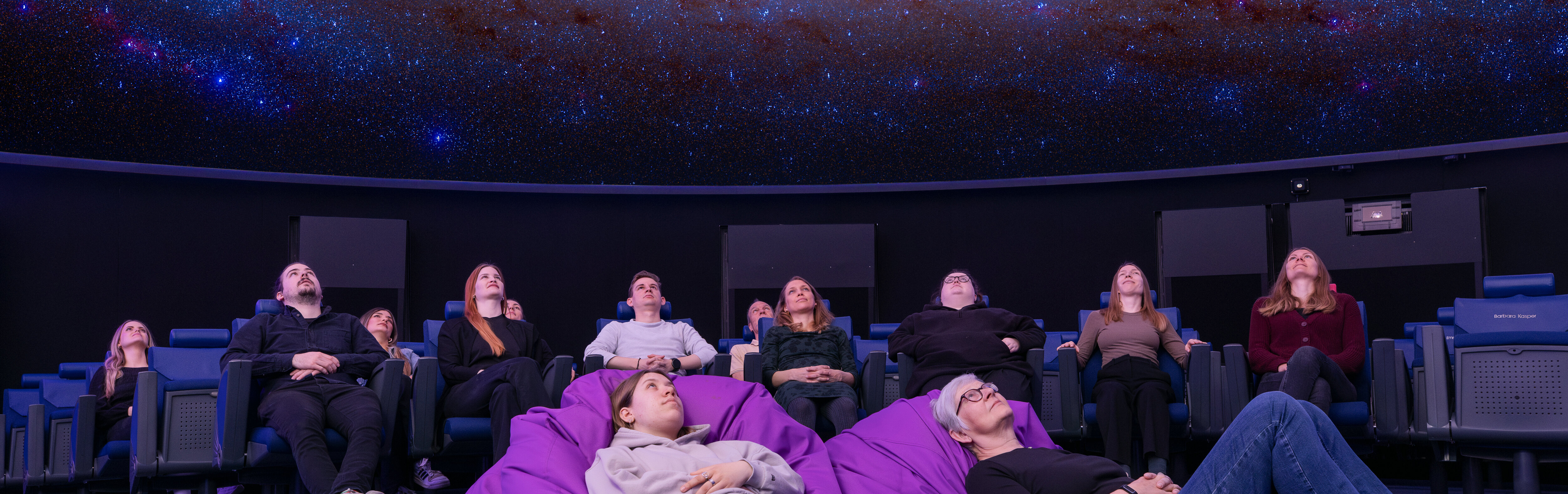 Personen sitzen in Lehnstühlen oder liegen in Sitzsäcken im Verkehrshaus Planetarium und schauen das Weltall an, das in der Kuppel projiziert wird. 