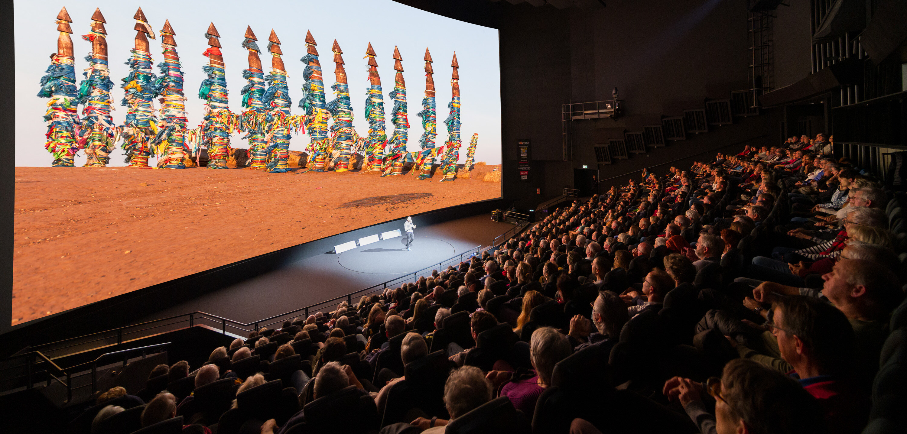 Ein modernes, gefülltes Kino mit einer grossen Projektionsleinwand, auf der eine Grafik mit farbigen Figuren zu sehen ist.