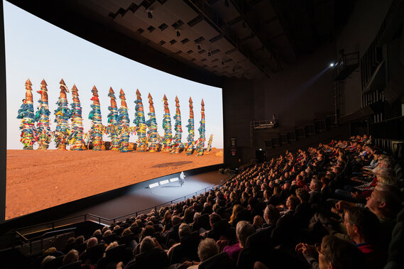 Ein modernes, gefülltes Kino mit einer grossen Projektionsleinwand, auf der eine Grafik mit farbigen Figuren zu sehen ist. 