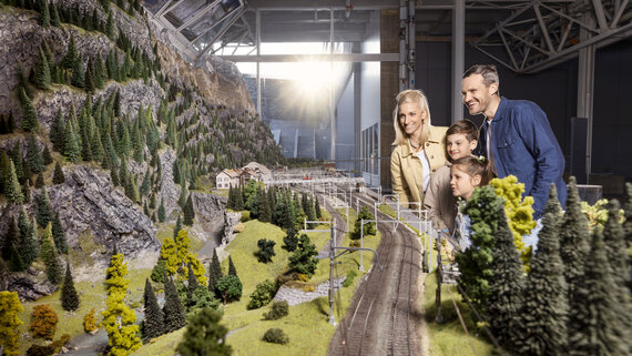 Eine vierköpfige Familie steht am Gotthardbahnmodell und betrachtet die Modelllandschaft. 