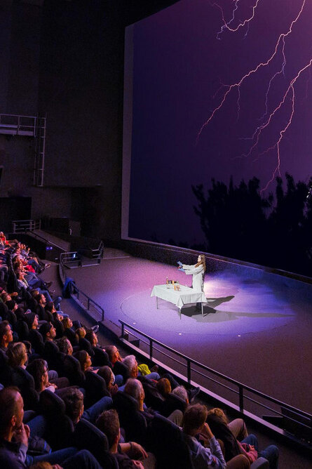 Ein gefüllter Filmtheatersaal im Verkehrshaus. Eine Frau im weissen Kittel steht auf der Bühne und präsentiert ein Experiment. 
