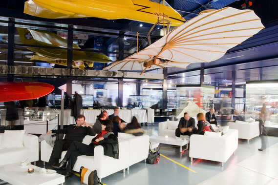 Personen sitzen auf weissen Lounge Sofas unter hängenden Fliegern in einer Halle.