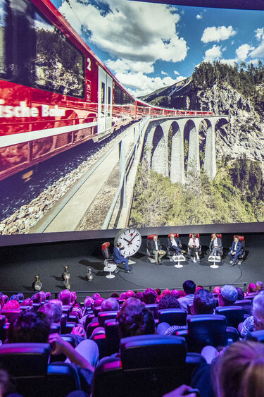 Ein gefüllter Kinosaal, vor der Leinwand sitzen Personen mit Stühlen und diskutieren, auf der Leinwand ist eine Eisenbahn zu sehen.