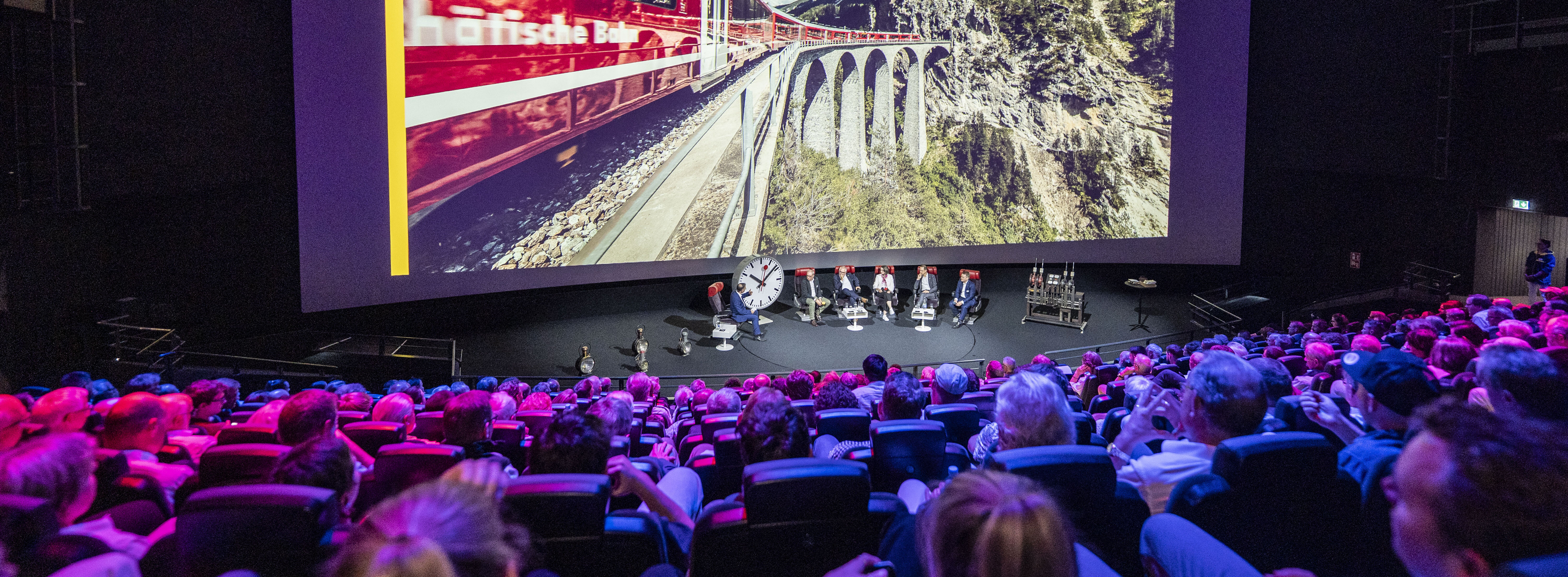 Ein gefüllter Kinosaal, vor der Leinwand sitzen Personen mit Stühlen und diskutieren, auf der Leinwand ist eine Eisenbahn zu sehen.