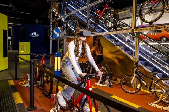 Ein Radfahrerin übt drinnen auf einem stationären Fahrrad mit einer VR-Brille, während an den Wänden verschiedene Fahrräder in einer lebendigen Museumsumgebung ausgestellt sind.