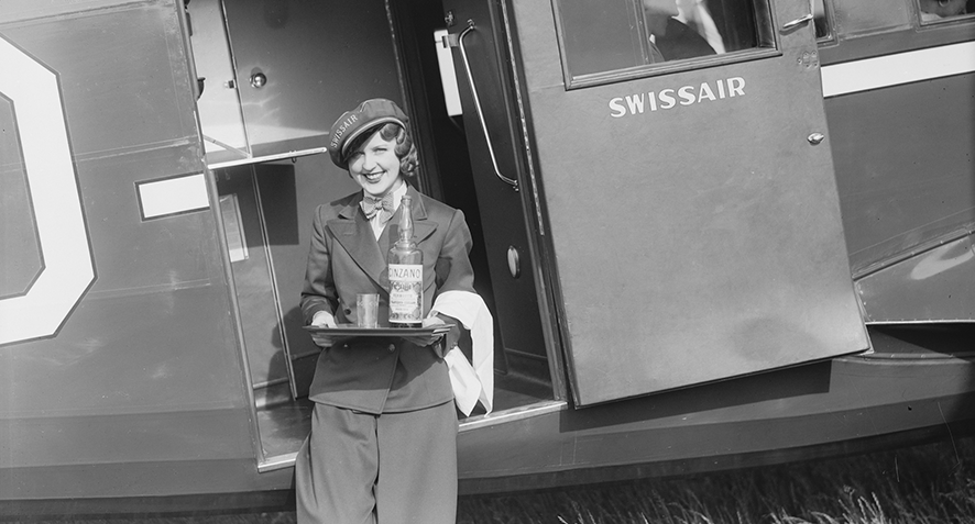 Eine Flugbegleiterin in Uniform hält ein Tablett mit einer Flasche und einem Glas in der Hand, während sie an der offenen Tür eines alten Swissair-Flugzeugs steht.