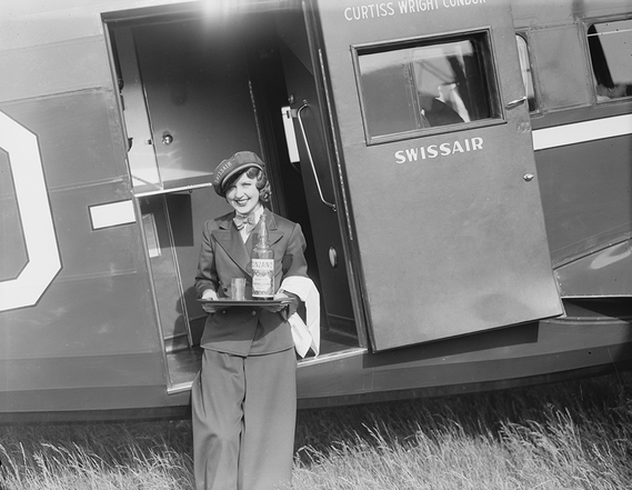 Eine Flugbegleiterin in Uniform hält ein Tablett mit einer Flasche und einem Glas in der Hand, während sie an der offenen Tür eines alten Swissair-Flugzeugs steht.