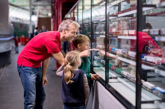 Eine Vater mit zwei Kinder schaut in einen Schaukasten mit Modelleisenbahnen und zeigt auf eine Bahn.