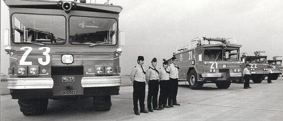 Eine Reihe alter Feuerwehrfahrzeuge mit den Nummern 21, 22 und 23, deren Uniformierte auf dem Rollfeld stramm stehen.