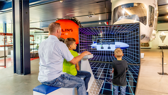 Eine Familie interagiert mit einer Flugsimulation in einem Museum, das von Flugzeugexponaten und einer lebendigen pädagogischen Atmosphäre umgeben ist.