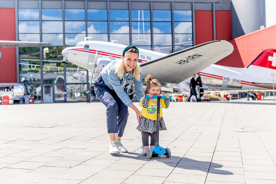  Ein Kind auf einem Motorroller, unterstützt von einem Erwachsenen, in der Nähe von alten Flugzeugen.