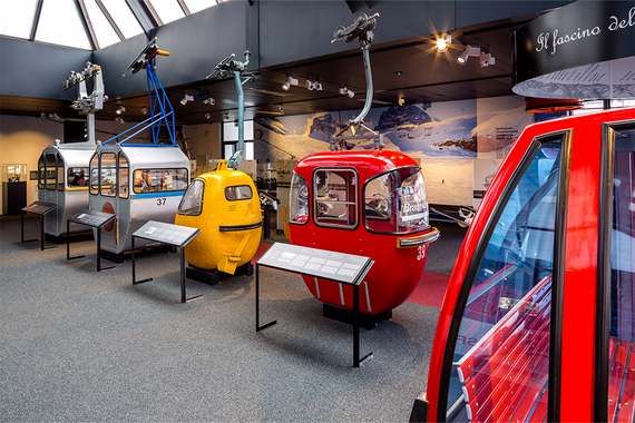 Vier alte Skigondeln in einer Museumsausstellung mit Modellen in Silber, Gelb und Rot vor einer malerischen Alpenkulisse.