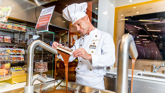 Ein Chocolatier in weisser Uniform träufelt gekonnt geschmolzene Schokolade aus einer Maschine, umgeben von verschiedenen Schokoladenprodukten.