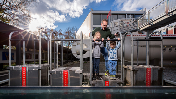 Zwei Kinder und ein Erwachsener stehen auf Plattformen an einem Wasserspiel, mit einem grossen Schiffsbauwerk im Hintergrund unter einem hellen Himmel.