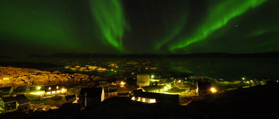 Grüne Polarlichter leuchten über einem kleinen Ort und dem Meer mit Eisschollen.