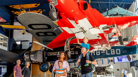 Eine Familie bewundert alte Flugzeuge, die in einem Museum ausgestellt sind. Über ihnen hängen bunte Flugzeuge in einer geräumigen, modernen Galerie.