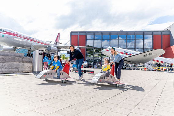 Zwei Kinder fahren fröhlich mit Miniaturflugzeugmodellen, während Erwachsene ihnen vor einem Luftfahrtmuseum mit grösseren Flugzeugen im Hintergrund helfen.
