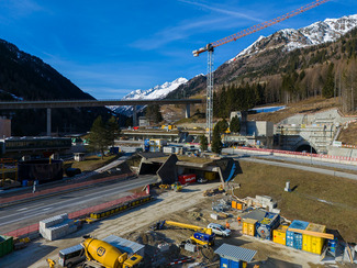 Eine Baustelle in bergigem Gelände mit Kränen, Betonstrukturen und Maschinen, umgeben von schneebedeckten Gipfeln.