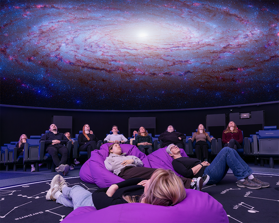 In einem Planetarium entspannen sich die Besucher auf Sitzsäcken und Stühlen unter einer anschaulichen Galaxienprojektion und blicken staunend nach oben.