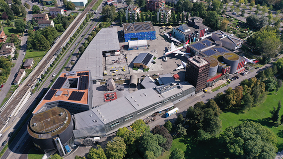 Luftaufnahme eines modernen Komplexes mit verschiedenen Gebäuden, einer Flugzeugausstellung und nahegelegenen Bahngleisen, umgeben von Grünflächen.