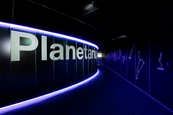 Ein geschwungener Korridor, der von violetten Lichtern beleuchtet wird, zeigt grosse „Planetarium“-Schilder und Umrisse von Sternbildern an dunklen Wänden.