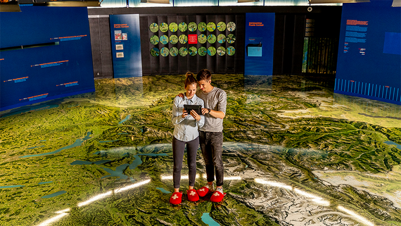 Ein Paar steht auf einer topografischen 3D-Karte, beschäftigt mit einem interaktiven Gerät, umgeben von bunten, informativen Displays.