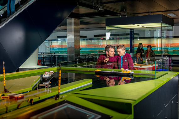 Zwei Kinder beobachten aufmerksam ein Schiffsmodell in einer Glasvitrine, umgeben von farbenfrohen, interaktiven Exponaten in einem Museum.
