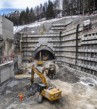 Eine Baustelle mit schweren Maschinen, darunter Bagger, die an einem Tunneleingang arbeiten, umgeben von felsigem Gelände und Bäumen.