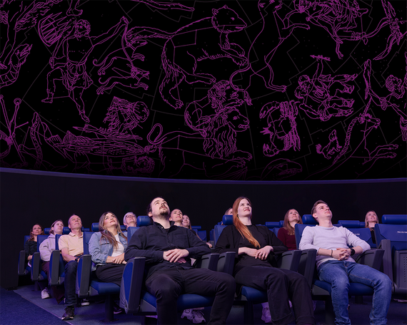 Die Zuschauer sitzen in einem Planetarium und blicken auf eine farbenfrohe Decke, an der die Sternbilder in Rosa vor einem dunklen Hintergrund abgebildet sind.