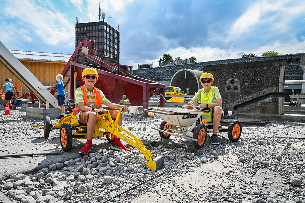 Zwei Kinder in leuchtenden Sicherheitswesten sitzen auf Mini-Spielzeug-Baufahrzeugen, umgeben von Kies und Baumaschinen.