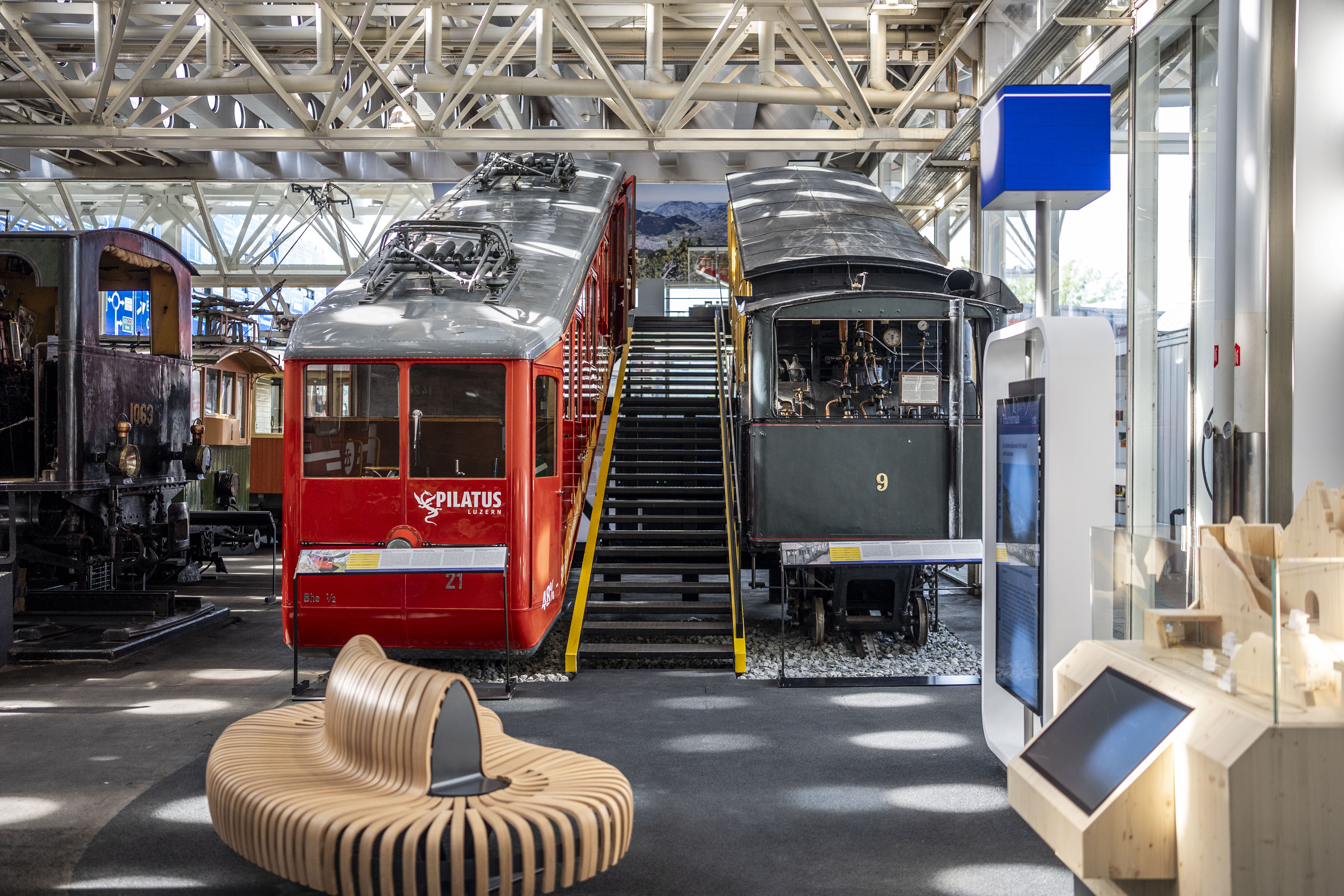 Zwei Zahnradbahnen stehen schräg nebeneinander, dazwischen führt eine Treppe hoch. Davor ist eine Holzbank und zwei Bildschirme zu sehen.