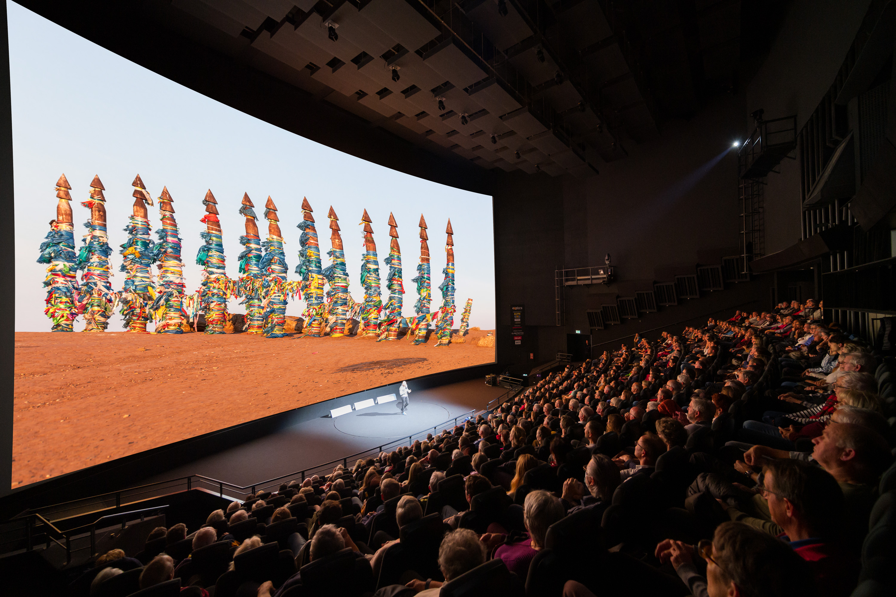 Ein modernes, gefülltes Kino mit einer grossen Projektionsleinwand, auf der eine Grafik mit farbigen Figuren zu sehen ist.