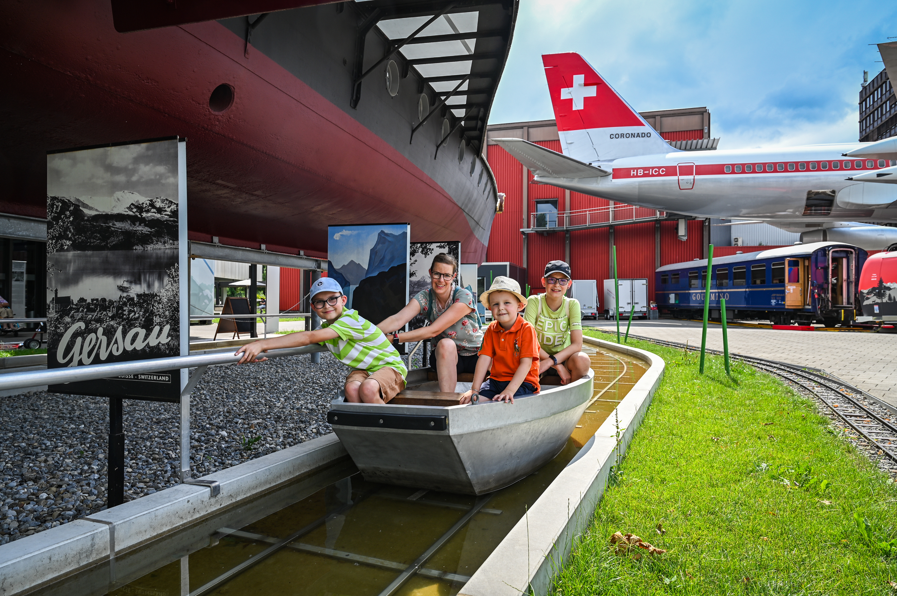 Drei Kinder und eine erwachsene Person sitzen in einem Ruderboot in einem Kanal. Hinter Ihnen ist ein Flugzeug zu sehen.