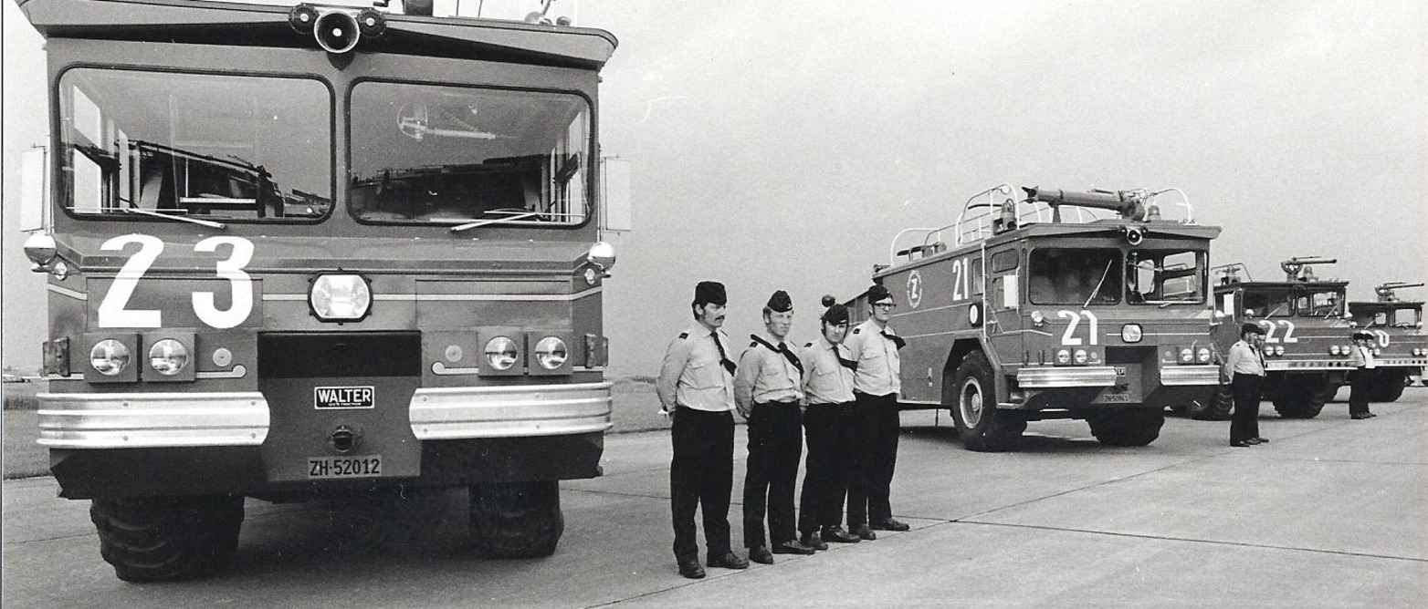 Eine Reihe alter Feuerwehrfahrzeuge mit den Nummern 21, 22 und 23, deren Uniformierte auf dem Rollfeld stramm stehen.